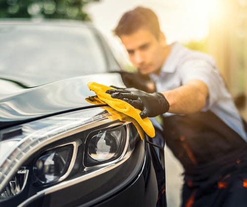 A man cleaning the headlight of a black car with a yellow microfiber cloth, illustrating car maintenance and detailing.