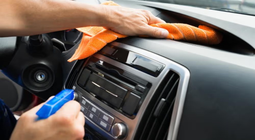 A person cleaning a car dashboard with an orange cloth and spray bottle.