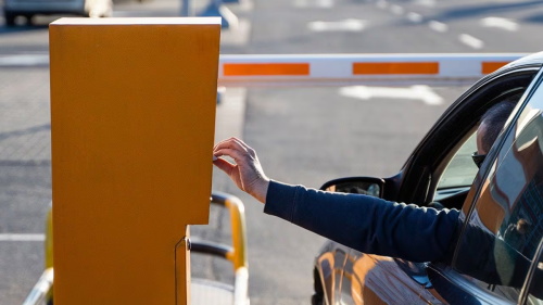 A driver reaching out of their car window to use a parking gate control.