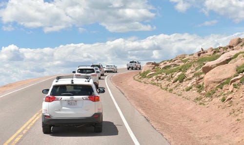 A line of white SUVs driving up a winding mountain road under a blue sky.
