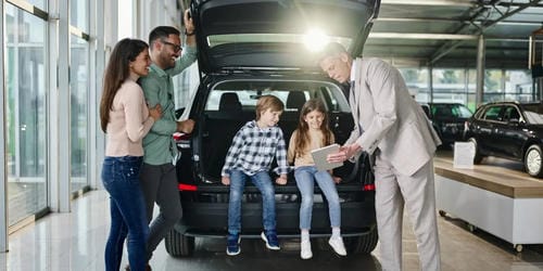 A family with two children at a car dealership, looking at the trunk space of an SUV with a salesperson.