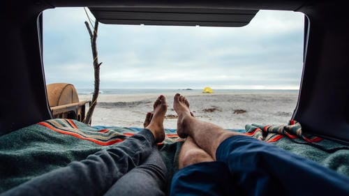 A couple relaxing inside the back of a vehicle parked on a beach, with their feet up and the ocean view ahead.
