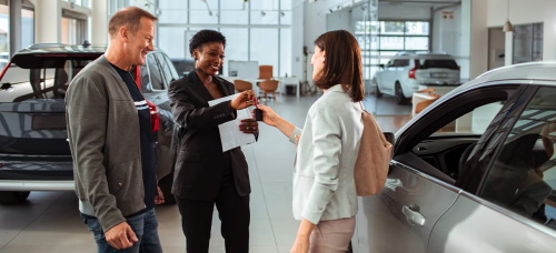A salesperson handing over car keys to a couple in a car dealership showroom.