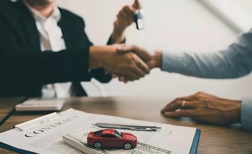 A handshake between buyer and seller over a contract, with a toy car and cash on the table.