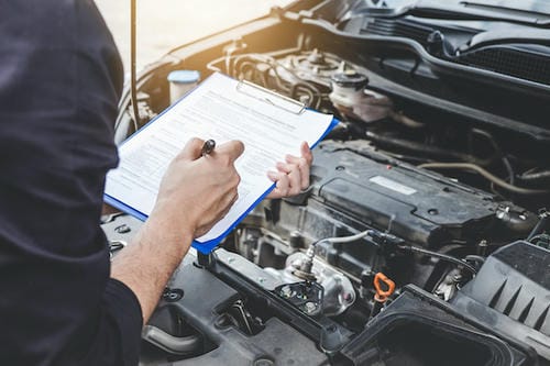 A mechanic writing on a clipboard while inspecting the engine bay of a car, likely during a service or safety check.