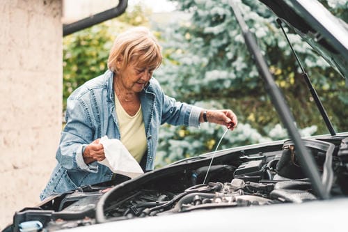 An older woman checking her car’s engine oil level using the dipstick, with the hood open.