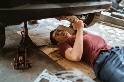 A mechanic working underneath a car using a wrench, with a car jack supporting the vehicle.