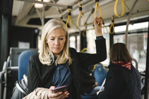 A woman using her phone while standing on a city bus, holding onto a hand strap.